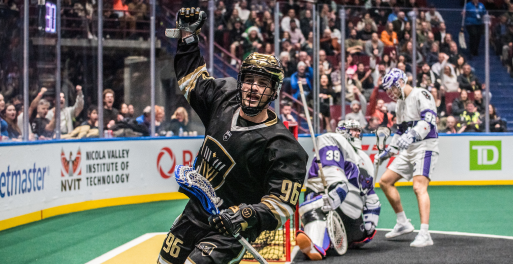 A lacrosse player in a black uniform raises his arm in celebration after scoring a goal, with fans cheering in the background. Two opposing players in white uniforms, including a goalie, react to the play.