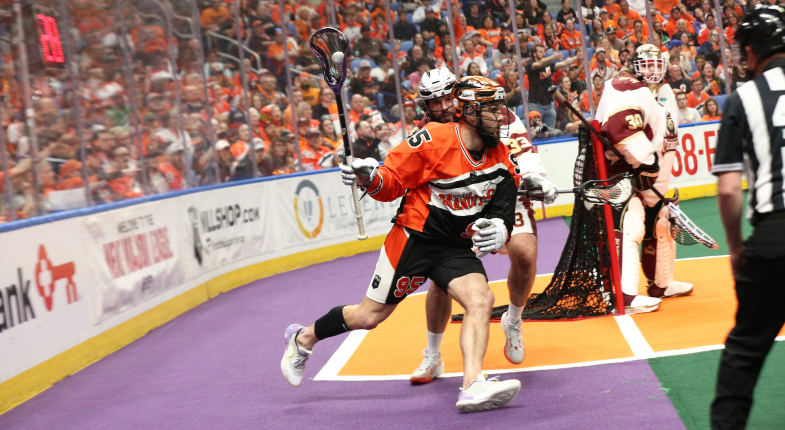 Lacrosse player in an orange jersey dodges defenders while aiming at the goal during a game.